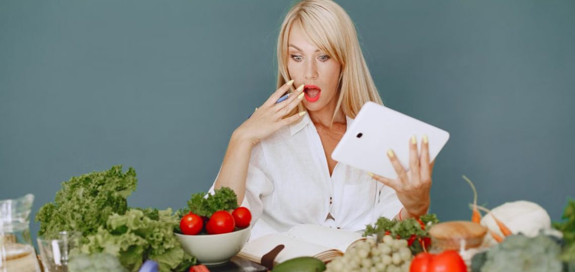 A surprised woman looking at a tablet surrounded by fresh vegetables and fruits, promoting healthy eating.